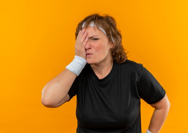 Спортивная женщина средних лет в черной футболке с повязкой на голове выглядит усталой, прикрывая один глаз рукой, стоящей над оранжевой стеной