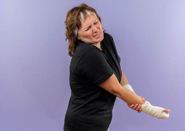 Middle aged sporty woman in black t-shirt with headband looking unwell holding her bandaged wrist having pain standing over blue wall