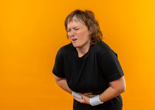 Спортивная женщина средних лет в черной футболке с повязкой на голове выглядит нездоровой, касаясь своего живота, страдающего от боли, стоя над оранжевой стеной