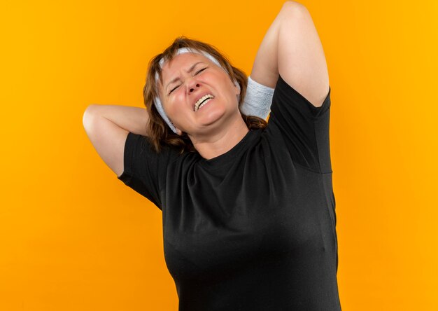 Free photo middle aged sporty woman in black t-shirt with headband looking unwell touching her neck having pain standing over orange wall