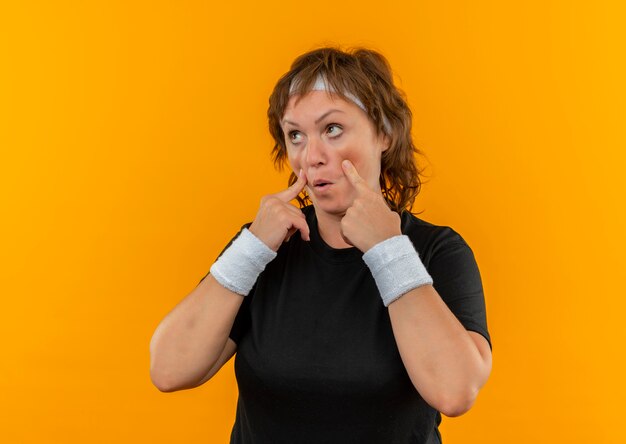 Middle aged sporty woman in black t-shirt with headband pointing with fingers ti her eyes watching you gesture standing over orange wall