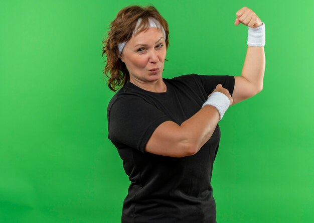 Middle aged sporty woman in black t-shirt with headband raising fist showing biceps looking confident and happy standing over green wall