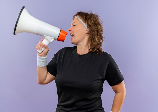Middle aged sporty woman in black t-shirt with headband shouting to megaphone standing over blue wall