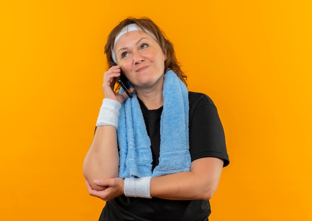 Спортивная женщина средних лет в черной футболке с повязкой на голову и полотенцем на плече, глядя в сторону, счастливая и позитивная, разговаривает по мобильному телефону, стоя над оранжевой стеной