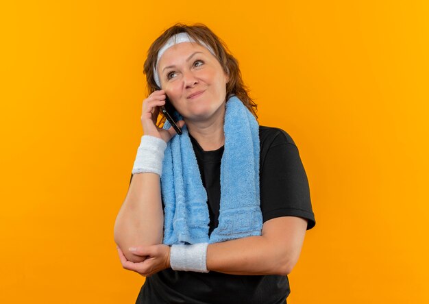 Middle aged sporty woman in black t-shirt with headband and towel on shoulder looking aside happy and positive, talking on mobile phone standing over orange wall