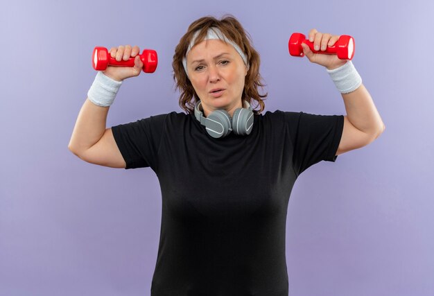 Спортивная женщина средних лет в черной футболке с повязкой на голове тренируется с гантелями, выглядит усталой и измученной, стоя у синей стены