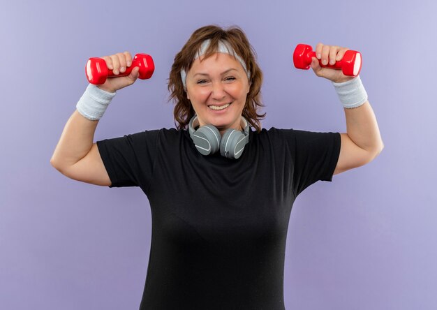 Middle aged sporty woman in black t-shirt with headband working out with dumbbells smiling cheerfully standing over blue wall