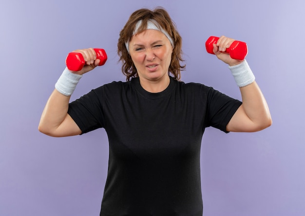 Спортивная женщина средних лет в черной футболке с повязкой на голове тренируется с двумя гантелями, усталая, стоя у синей стены