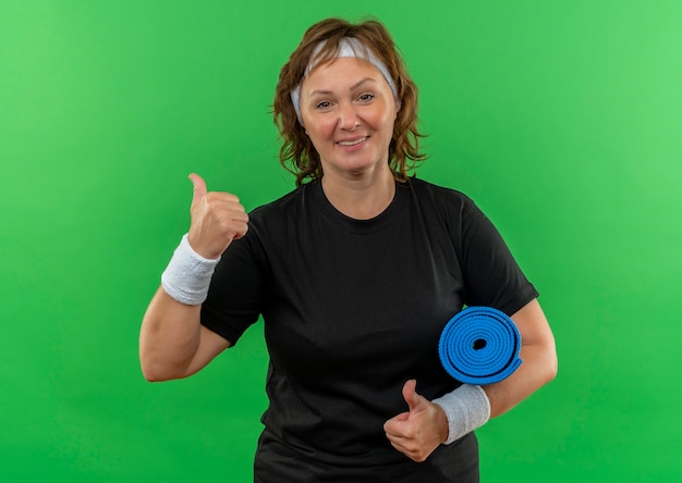 Бесплатное фото Спортивная женщина средних лет в черной футболке с повязкой на голове держит коврик для йоги, весело улыбаясь, показывает палец вверх, стоя над зеленой стеной