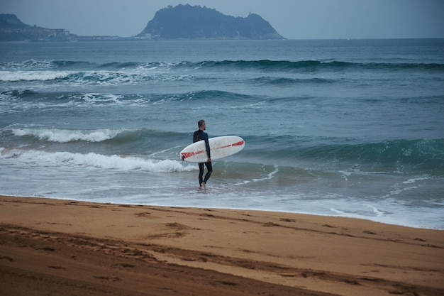 Middle aged surfer in wetsuit walking into the water on sandy beach among hills