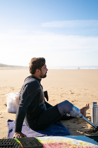 Middle-aged surfer with artificial leg sitting on beach and looking at sea