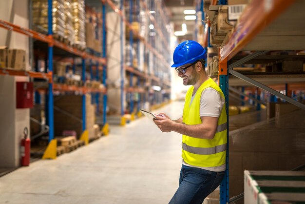 Middle aged warehouse worker with hardhat using tablet in large storehouse distribution area