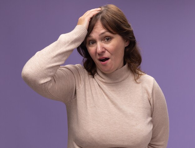 Middle aged woman in beige turtleneck  confused and very anxious with hand on her head for mistake standing over purple wall