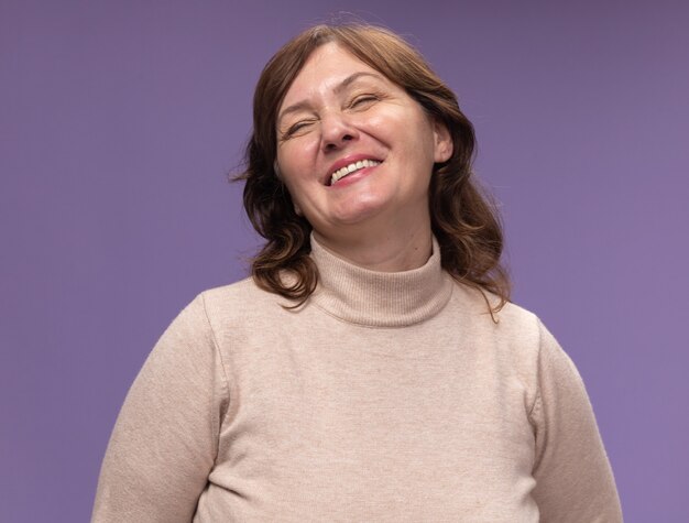 Middle aged woman in beige turtleneck happy and cheerful smiling with closed eyes standing over purple wall