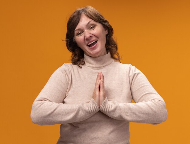 Middle aged woman in beige turtleneck holding hands together like namaste smiling friendly standing over orange wall
