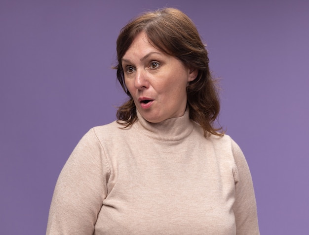 Free photo middle aged woman in beige turtleneck looking aside confused and surprised standing over purple wall