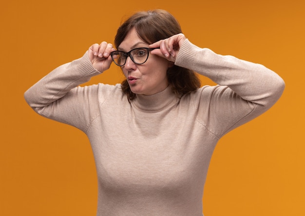 Donna di mezza età in dolcevita beige con gli occhiali che osserva da parte da vicino in piedi sopra la parete arancione