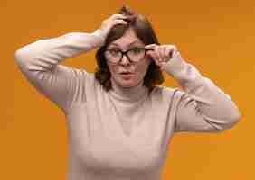 Free photo middle aged woman in beige turtleneck wearing glasses  surprised standing over orange wall