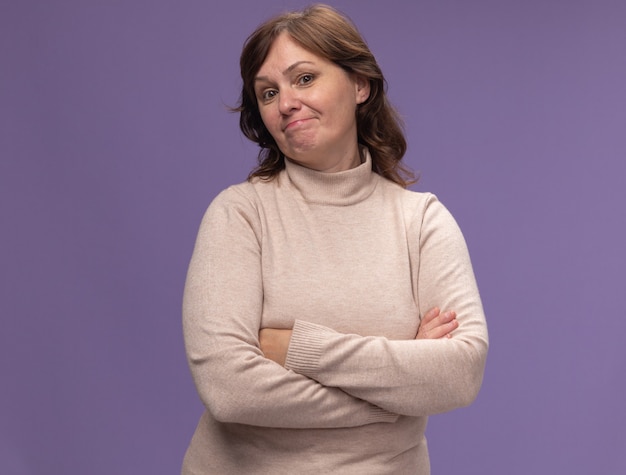 Middle aged woman in beige turtleneck  with skeptic expression with arms crossed standing over purple wall