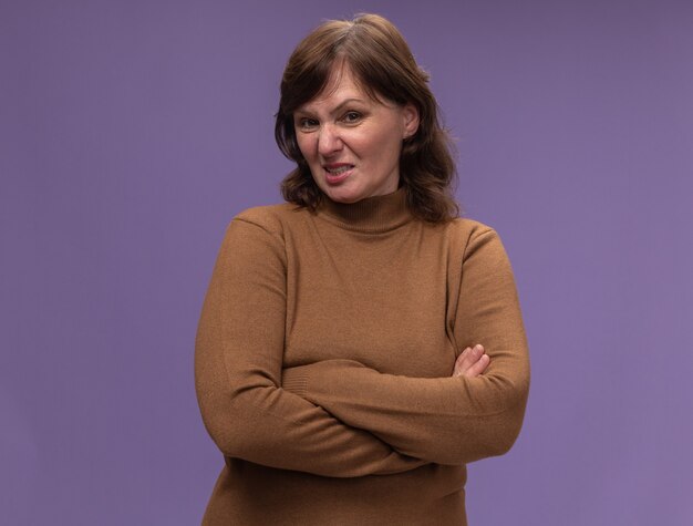 Middle aged woman in brown turtleneck  baing annoyed and irritated with arms crossed standing over purple wall