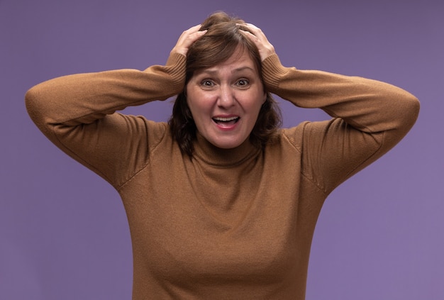 Free photo middle aged woman in brown turtleneck  confused with hands on her head for mistake standing over purple wall