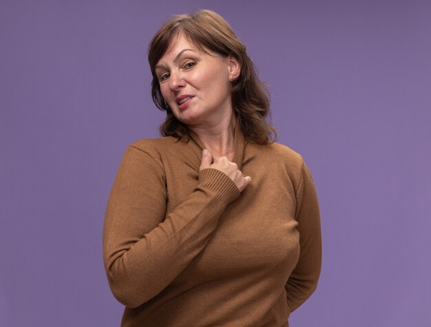 Женщина средних лет в коричневой водолазке выглядит раздраженной и раздраженной, касаясь своей шеи, стоящей над фиолетовой стеной