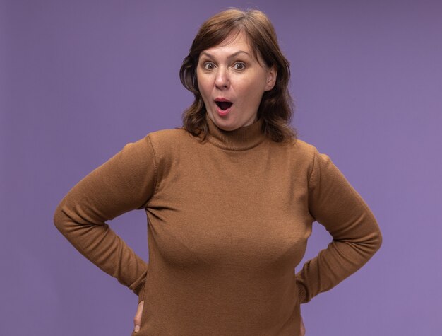 Free photo middle aged woman in brown turtleneck standing over purple wall