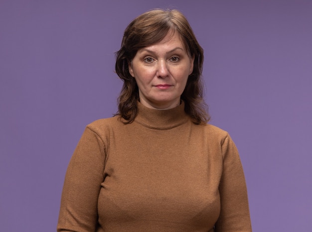 Free photo middle aged woman in brown turtleneck  with confident expression standing over purple wall