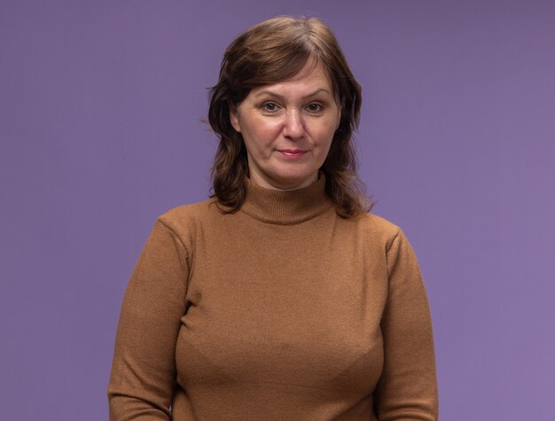 Женщина средних лет в коричневой водолазке со скептическим выражением лица стоит у фиолетовой стены