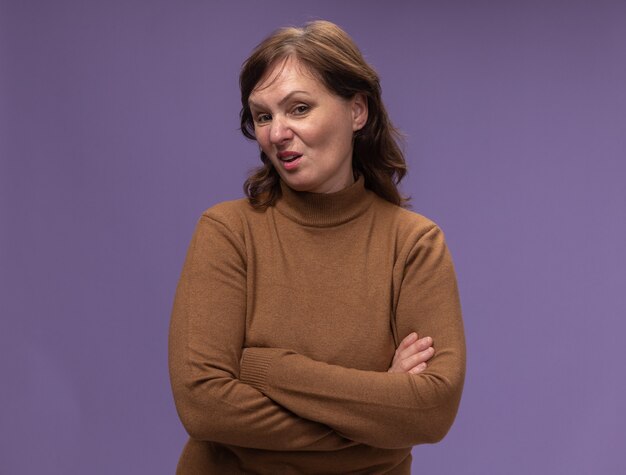 Middle aged woman in brown turtleneck  with skeptic expression with arms crossed standing over purple wall