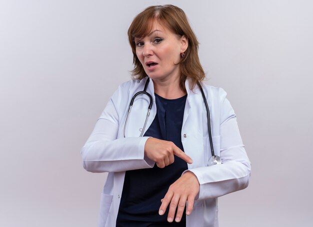 中年の女性医師が医療用ローブとコピースペースと孤立した白い壁に彼女の手で指で指している聴診器を着ています。