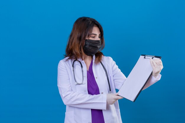 Middle aged woman doctor wearing white coat in black protective facial mask and with stethoscope holding clipboard looking at it with serious and confident expression on face standing over blue