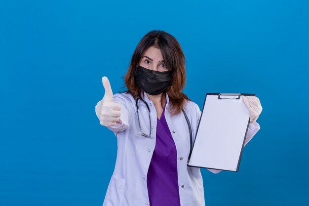 Middle aged woman doctor wearing white coat in black protective facial mask and with stethoscope holding clipboard with blanks looking positive showing thumbs up standing over blue background