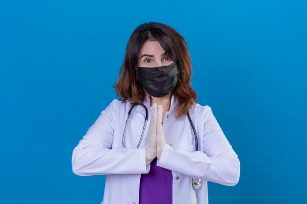 Женщина-врач средних лет в белом халате в черной защитной маске для лица и со стетоскопом, держась за руки в молитвенном жесте намасте, чувствуя благодарность и счастье на изолированном синем фоне