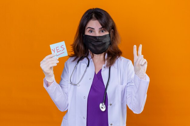 Middle aged woman doctor wearing white coat in black protective facial mask and with stethoscope holding reminder paper with yes word showing victory sign over orange background