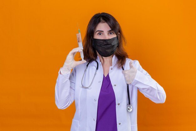 中年の女性医師が黒い保護用の顔のマスクに白のコートを着て、オレンジ色のれたらの上に立ってカメラを見て親指を示す陽気で幸せな親指を見て注射器を保持している聴診器で