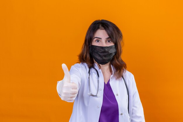 Middle aged woman doctor wearing white coat in black protective facial mask and with stethoscope looking at camera positive and happy showing thumbs up standing over orange background