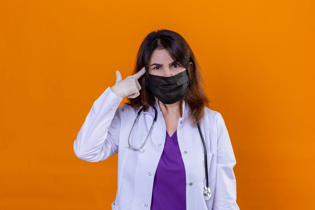 Middle aged woman doctor wearing white coat in black protective facial mask and with stethoscope pointing temple with finger concentrating hard on an idea with a serious look standing over iso