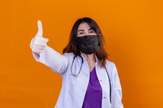 Женщина-врач средних лет в белом халате в черной защитной маске для лица и со стетоскопом положительно и счастливо смотрит в камеру, показывая большие пальцы руки вверх, стоя на оранжевом фоне