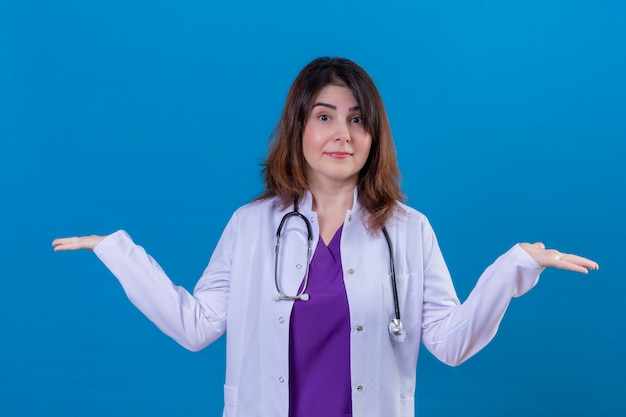 Женщина-врач средних лет в белом халате и со стетоскопом, невежественная и смущенная с распростертыми объятиями, понятия не имею, стоит на изолированном синем фоне