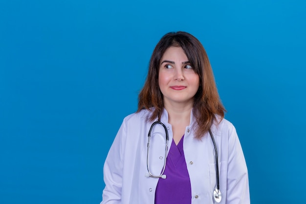 Middle aged woman doctor wearing white coat and with stethoscope looking aside smiling slyly standing over blue background
