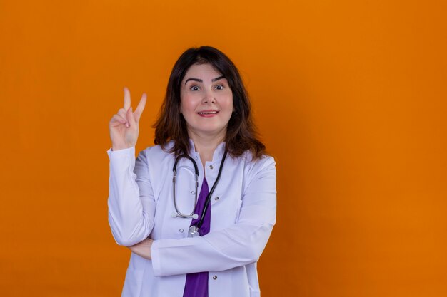 Middle aged woman doctor wearing white coat and with stethoscope looking at camera positive and happy showing victory sign smiling standing over isolated orange background