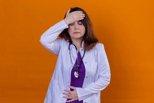 Middle aged woman doctor wearing white coat and with stethoscope looking unwell tired and overworked touching head having headache standing over isolated orange background