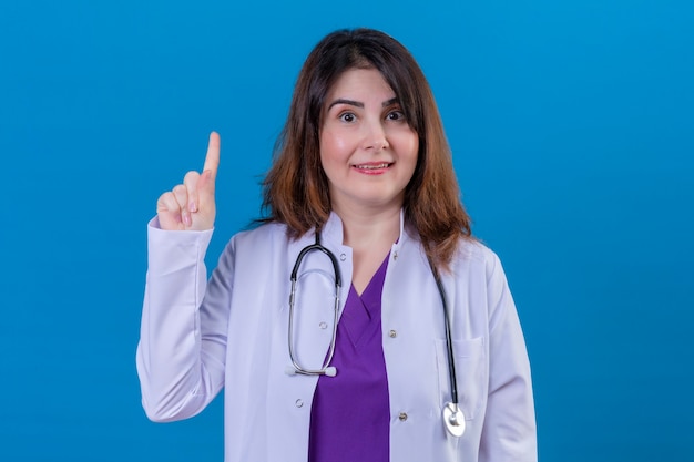 Женщина-врач средних лет в белом халате и со стетоскопом, указывающим вверх с улыбающимся пальцем, уверенно сосредоточилась на задаче, стоящей на изолированном синем фоне