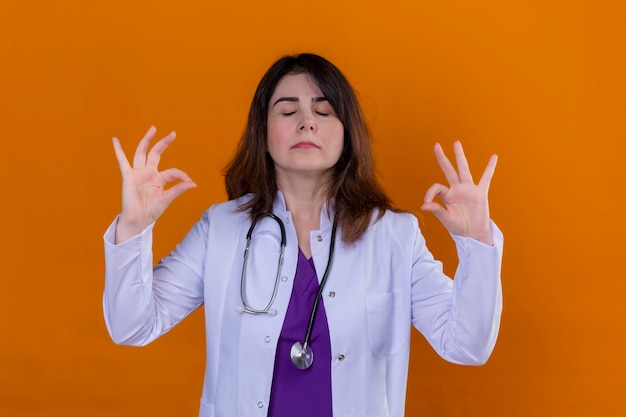 Foto gratuita medico donna di mezza età che indossa camice bianco e con lo stetoscopio rilassarsi e sorridere con gli occhi chiusi facendo il gesto di meditazione con le dita su sfondo arancione