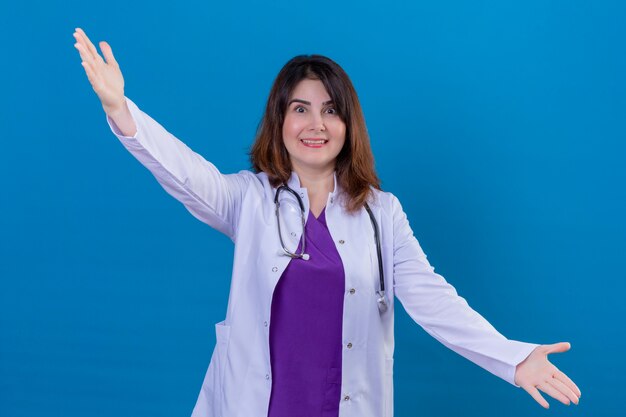 Middle aged woman doctor wearing white coat and with stethoscope smiling making a welcome gesture with raised hands over blue background