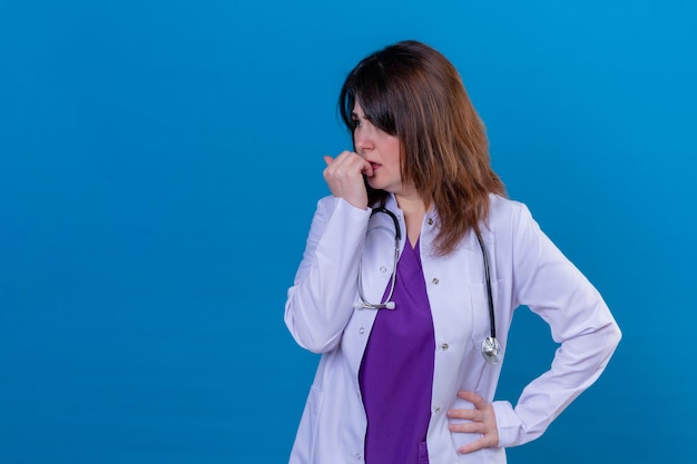 Middle aged woman doctor wearing white coat and with stethoscope stressed and nervous biting nails standing over blue background