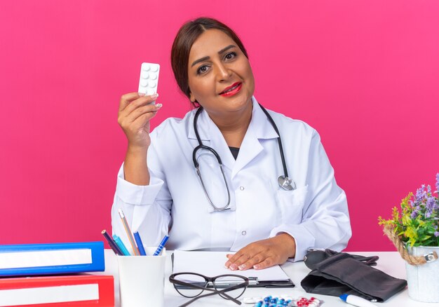 Middle aged woman doctor in white coat with stethoscope holding blister with pills looking at front smiling confident sitting at the table with office folders over pink wall