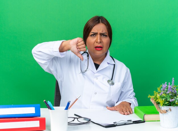 Женщина-врач средних лет в белом халате со стетоскопом, недовольно смотрящая на фронт, показывает палец вниз, сидя за столом над зеленой стеной