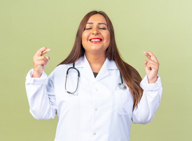 Middle aged woman doctor in white coat with stethoscope making desirable wish with smile on face crossing fingers standing over green wall